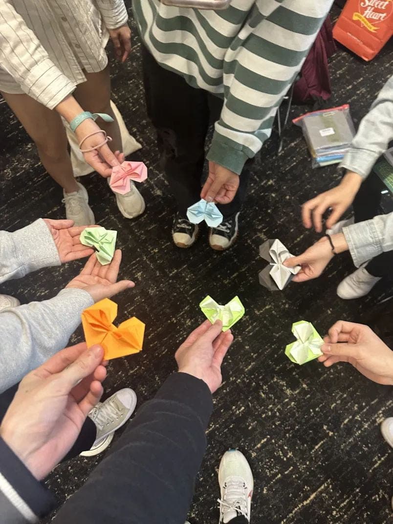 A circle of hands each holding a folded paper origami heart in a different colour.