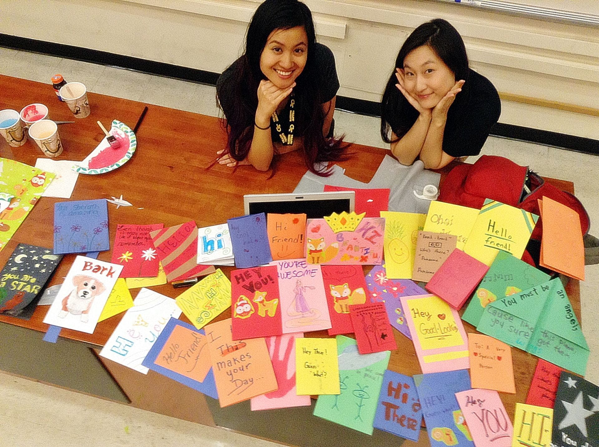Two members resting their chins on their hands behind a table covered in dozens of handmade greeting cards.